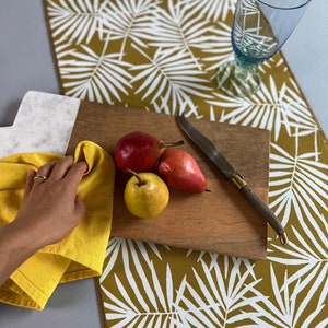 May include: A wooden cutting board with three pears and a knife rests on a mustard-colored table runner with a white palm leaf pattern. A yellow cloth and a blue glass are also present.