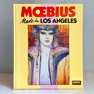 May include: A hardback book titled "Moebius Made in Los Angeles" with a yellow cover. The cover features an illustration of a woman with blue and purple accents. The book's title is in red and black text.