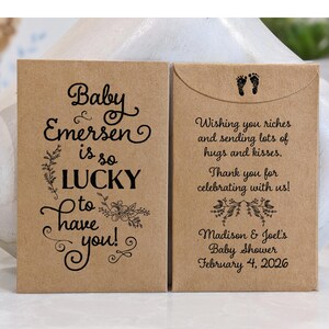 May include: Two brown paper favor bags with black text and floral designs. The left bag says "Baby Emersen is so LUCKY to have you!" The right bag says "Wishing you riches and sending lots of hugs and kisses. Thank you for celebrating with us! Madison & Joel's Baby Shower February 4, 2026"