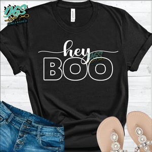 Könnte beinhalten: Schwarzes T-Shirt mit weißem Text, der "hey boo" lautet. Das Shirt liegt auf einer Holzoberfläche mit einer blauen Jeans und einem Paar Sandalen.