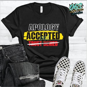May include: Black t-shirt with the text "Apology Accepted Trust Denied" in yellow and red lettering. The text is crossed out with a red line.