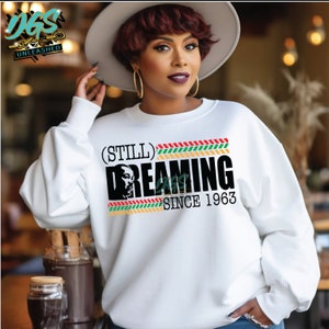 May include: White sweatshirt featuring the text "(Still) Dreaming Since 1963" in black, with a graphic of a person's face. The design includes a colourful stripe pattern. The sweatshirt has long sleeves.
