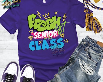 May include: A purple t-shirt with the text "Fresh est Senior in the Class" in a graffiti-style design. The shirt is paired with denim shorts and purple and white trainers. A graduation tassel is visible.
