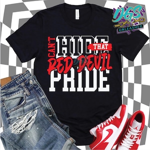 Puede incluir: Camiseta negra con el texto "Can't Hide That Red Devil Pride" en blanco y rojo. La imagen incluye vaqueros azules y zapatillas rojas y blancas, todo sobre un fondo a cuadros.