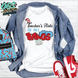 May include: White t-shirt featuring a red and blue graphic that reads "Teacher's Aide of All Things" with a cartoon cat wearing a red and white hat.
