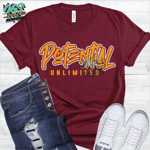 Könnte beinhalten: Ein maroonfarbenes T-Shirt mit dem Text "Potential Unlimited" in orangefarbener und weißer Schrift. Das Shirt ist auf einer Holzoberfläche mit einem Paar weißen Sneakers und einer blauen Jeans abgebildet.