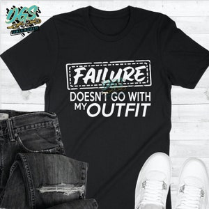 May include: Black t-shirt with white text that reads "FAILURE DOESN'T GO WITH MY OUTFIT". The text is in a box with a dashed border. The shirt is being modeled with jeans and white sneakers.