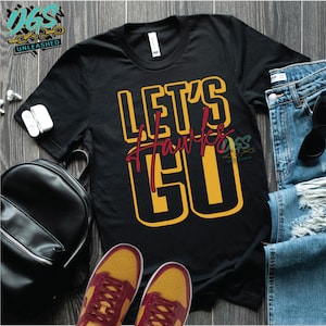 May include: Black t-shirt with the text "Let's Hawks Go" in yellow and red lettering. The shirt is laid out on a wooden surface with a pair of yellow and red sneakers and a black backpack.