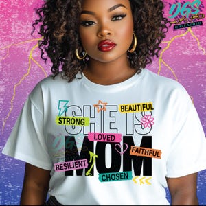 May include: A white t-shirt with the text "She is Mom" in black with colorful words and designs around it. The words include "Strong", "Loved", "Beautiful", "Faithful", "Resilient", and "Chosen".