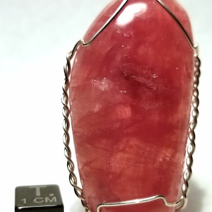 May include: A polished, oval-shaped pink gemstone pendant, encased in a silver wire frame. The stone has a translucent quality, with variations in color. A small black cube with "1 CM" is next to the pendant.