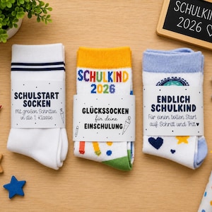 Könnte beinhalten: Drei Paar gefaltete Socken mit Papierbanderolen. Das linke Paar ist weiß mit blauen Streifen. Das mittlere Paar ist weiß mit gelbem Bündchen und bunten Schulmotiven. Das rechte Paar ist weiß mit hellblauem Bündchen und Sternenmustern.