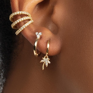 Peut inclure: Gros plan sur une oreille ornée de bijoux en or. L'oreille est ornée d'un ear cuff à trois rangs, d'une petite boucle d'oreille et d'une créole avec un charm en forme de palmier. Toutes les pièces sont en or et ornées de petites pierres claires.