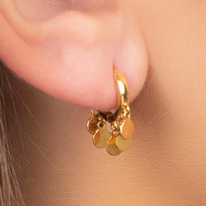 Può includere: Primo piano di un orecchino a cerchio dorato con piccoli ciondoli rotondi dorati pendenti. L'orecchino è attaccato a un orecchio. L'orecchino è un design semplice ed elegante.