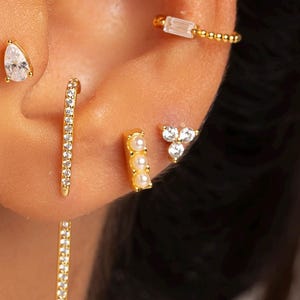 May include: A close-up of an ear adorned with multiple gold earrings. The earrings include a teardrop stud, a long bar with dangling crystals, a small bar with crystals, a triple-crystal stud, and a cuff earring with a rectangular crystal.