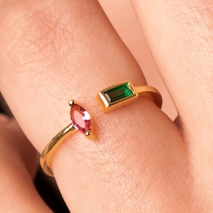 Peut inclure: Une bague dorée avec un design ouvert, ornée d'une pierre rose taillée en marquise et d'une pierre verte taillée en émeraude. La bague est portée sur un doigt, mettant en valeur les couleurs contrastées des pierres.