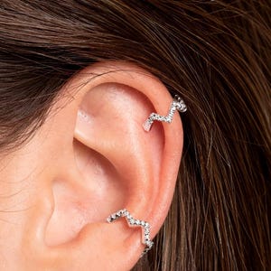 Könnte beinhalten: Nahaufnahme eines Ohrs mit silbernen Ohrringen. Ein kleiner, polierter Creolen-Ohrring befindet sich im Ohrläppchen, und zwei passende, wellenförmige Ohrklemmen mit kleinen, funkelnden Steinen befinden sich am oberen Ohrknorpel. Der Schmuck ist vor einem neutralen Hintergrund platziert.