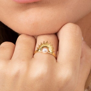 Può includere: Un anello d'oro con una perla al centro e un design a raggiera. L'anello è indossato al dito di una persona.