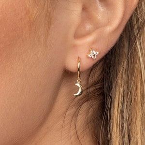 Può includere: Una donna indossa orecchini a cerchio dorati con un ciondolo a forma di luna crescente e un piccolo orecchino a perno dorato con un gruppo di pietre di zircone cubico trasparenti.