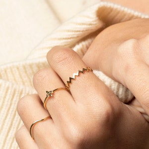 Può includere: Una mano con diversi anelli d'oro, tra cui una fascia a zig-zag, una fascia sottile con una piccola pietra preziosa e una fascia doppia.