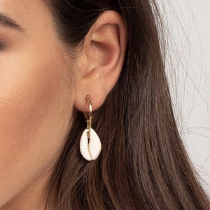 Puede incluir: Un par de pendientes de aro dorados con un dije de concha blanca colgando de cada aro.