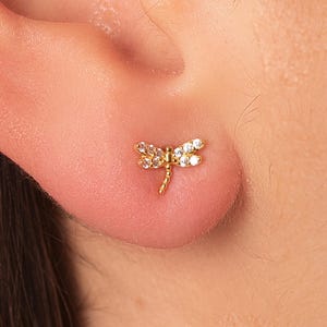 以下が含まれることがあります： 金色のトンボのスタッドピアスで、クリアでキラキラとしたアクセントが耳に表示されています。ピアスは小さく繊細なジュエリーで、トンボの羽と体がはっきりと定義されています。