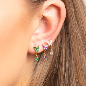 Può includere: Primo piano di un orecchio con tre orecchini pendenti. Gli orecchini sono dorati con ciondoli a forma di farfalla e pietre preziose di diversi colori. Gli orecchini sono verdi, viola e bianchi.