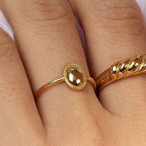 Könnte beinhalten: Zwei goldene Ringe an einem Finger. Ein Ring hat einen ovalen Mittelstein mit einem Perlenrand. Der andere Ring hat ein gedrehtes Seil-Design. Beide Ringe sind glänzend und reflektierend, vor weißem Hintergrund.