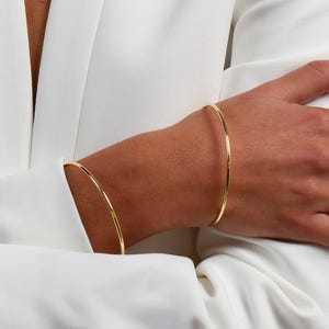 Può includere: Primo piano di una persona che indossa un blazer bianco e gioielli d'oro. I gioielli includono diversi anelli d'oro e due sottili braccialetti d'oro. È visibile anche un delicato braccialetto d'oro. Lo sfondo è di un colore neutro e morbido.