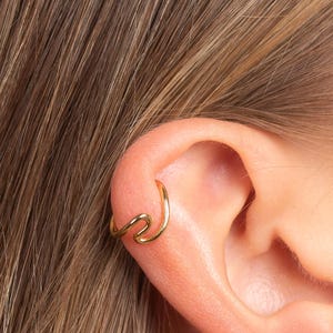 Könnte beinhalten: Ein goldfarbener Ohrklemme mit wellenförmigem Design ist an einem Ohr zu sehen. Der Ohrschmuck ist ein einfaches, elegantes Schmuckstück. Das Haar der Person ist hellbraun.