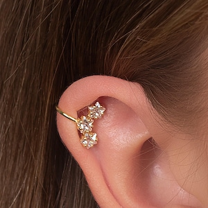 Può includere: Un orecchino a cerchio dorato con tre cristalli trasparenti a forma di stella. L'orecchino si curva attorno all'orecchio, con i cristalli raggruppati. Il gioiello è ambientato sullo sfondo di un orecchio e capelli castani.
