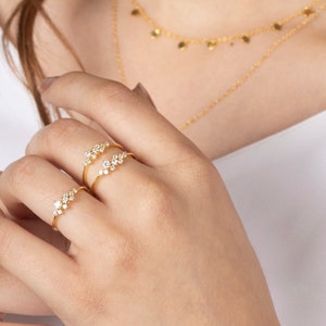 May include: Three gold rings with small, clear stones set in a cluster design. The rings are worn on the index finger of a hand.
