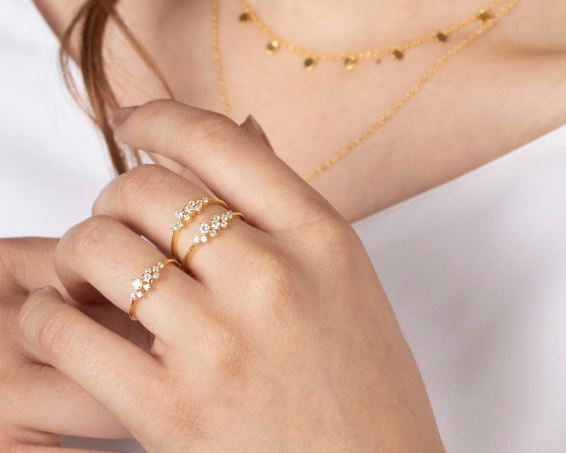 May include: Three gold rings with small, clear stones set in a cluster design. The rings are worn on the index finger of a hand.