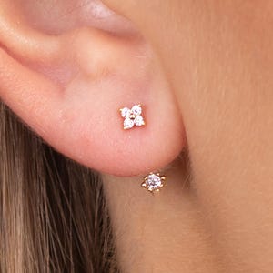 May include: Gold stud earrings. One earring has a four-petal flower design with clear stones. The other earring features a single, round clear stone. The earrings are set against the skin of the ear.