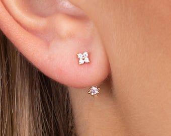 May include: Gold-toned stud earrings. One earring features a four-petal flower design with clear stones. The other earring has a single, round clear stone. The earrings are set against the skin of the ear.