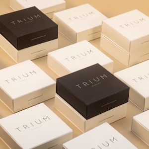 May include: A collection of black and white square boxes with the text 'TRIUM Jewelry' printed on them. The boxes are arranged in a grid pattern on a light brown surface.