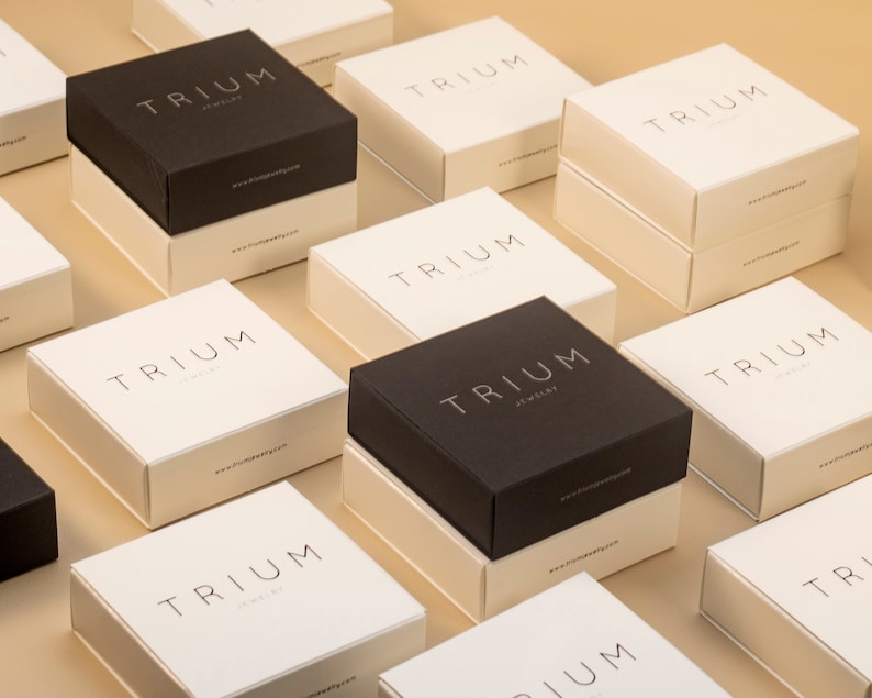 May include: A collection of black and white square boxes with the text 'TRIUM Jewelry' printed on them. The boxes are arranged in a grid pattern on a light brown surface.