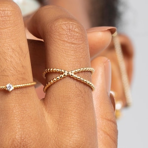 Könnte beinhalten: Nahaufnahme von drei goldenen Ringen. Ein Ring hat einen kleinen, klaren Edelstein. Ein weiterer Ring hat ein Kreuzmuster aus kleinen goldenen Perlen. Der dritte Ring ist ein schlichtes goldenes Band. Die Ringe befinden sich an den Fingern einer Person.