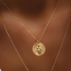 Könnte beinhalten: Goldfarbene Halsketten in mehreren Lagen. Die obere Halskette hat einen kreisförmigen Anhänger mit einem Sonnenstrahl-Design und einer Schlangensilhouette. Die zweite Halskette ist eine einfache Kette. Der Schmuck wird vor einem Hautton-Hintergrund getragen.