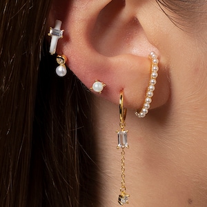 May include: A gold hoop earring with a dangling crystal charm. The hoop is adorned with small white pearls. The earring is paired with a white pearl stud earring and a white bar earring with a small crystal charm.