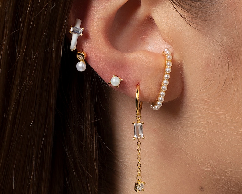 May include: A gold hoop earring with a dangling crystal charm. The hoop is adorned with small white pearls. The earring is paired with a white pearl stud earring and a white bar earring with a small crystal charm.