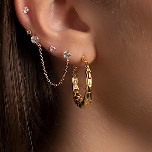 Peut inclure: Gros plan sur une oreille ornée de multiples boucles d'oreilles dorées. L'oreille présente de petites boucles d'oreilles à clous et une chaîne reliant deux boucles d'oreilles plus grandes et scintillantes. Une boucle d'oreille créole dorée texturée complète le look.