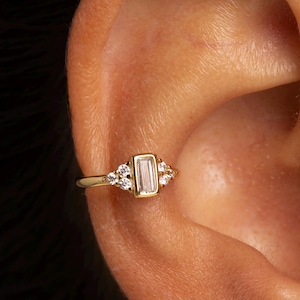 Può includere: Un orecchino a cerchio dorato con una pietra rettangolare di colore chiaro al centro, affiancata da pietre rotonde e trasparenti più piccole. Il cerchio è progettato per avvolgere la cartilagine dell'orecchio, creando un look delicato ed elegante. Il gioiello è su sfondo scuro.