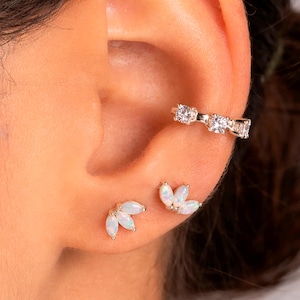 Può includere: Primo piano di un orecchio adornato da tre gioielli. Un ear cuff argentato con tre pietre trasparenti si trova sulla parte superiore dell'orecchio. Sono visibili anche due serie di piccoli orecchini a lobo in opale bianco.