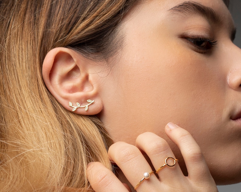 May include: A gold leaf ear cuff with small clear stones. The ear cuff is worn on the left ear of a person with long blonde hair. The person is also wearing a gold ring with a pearl on the ring finger of their right hand.