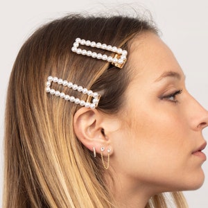 Puede incluir: Dos pinzas para el pelo doradas con filas de perlas blancas. Las pinzas est&aacute;n en el pelo de una mujer. Tambi&eacute;n lleva pendientes de oro con peque&ntilde;as piedras transparentes.