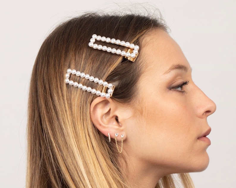 Puede incluir: Dos pinzas para el pelo doradas con filas de perlas blancas. Las pinzas est&aacute;n en el pelo de una mujer. Tambi&eacute;n lleva pendientes de oro con peque&ntilde;as piedras transparentes.