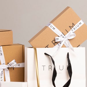 May include: A white shopping bag with black handles and the text "TRIUM JEWELRY" printed on the front. The bag is filled with brown cardboard boxes wrapped with white ribbon and the text "TRIUM" printed on the ribbon.