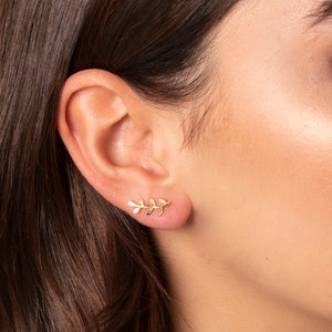 May include: A close-up of a person's ear with a gold leaf-shaped earring. The earring is a delicate piece of jewelry that would make a great gift for a friend or loved one.