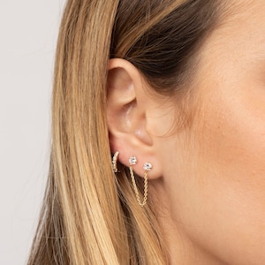 Puede incluir: Primer plano de la oreja de una persona con tres pendientes de oro. Los pendientes son un aro peque&ntilde;o con una fila de peque&ntilde;os diamantes, un peque&ntilde;o diamante de bot&oacute;n y un diamante de bot&oacute;n con una cadena colgante.