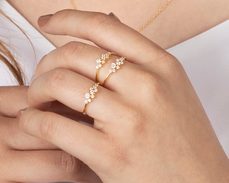 May include: Two gold rings with small, clear stones set in a cluster design. The rings are worn on the index finger of a hand.